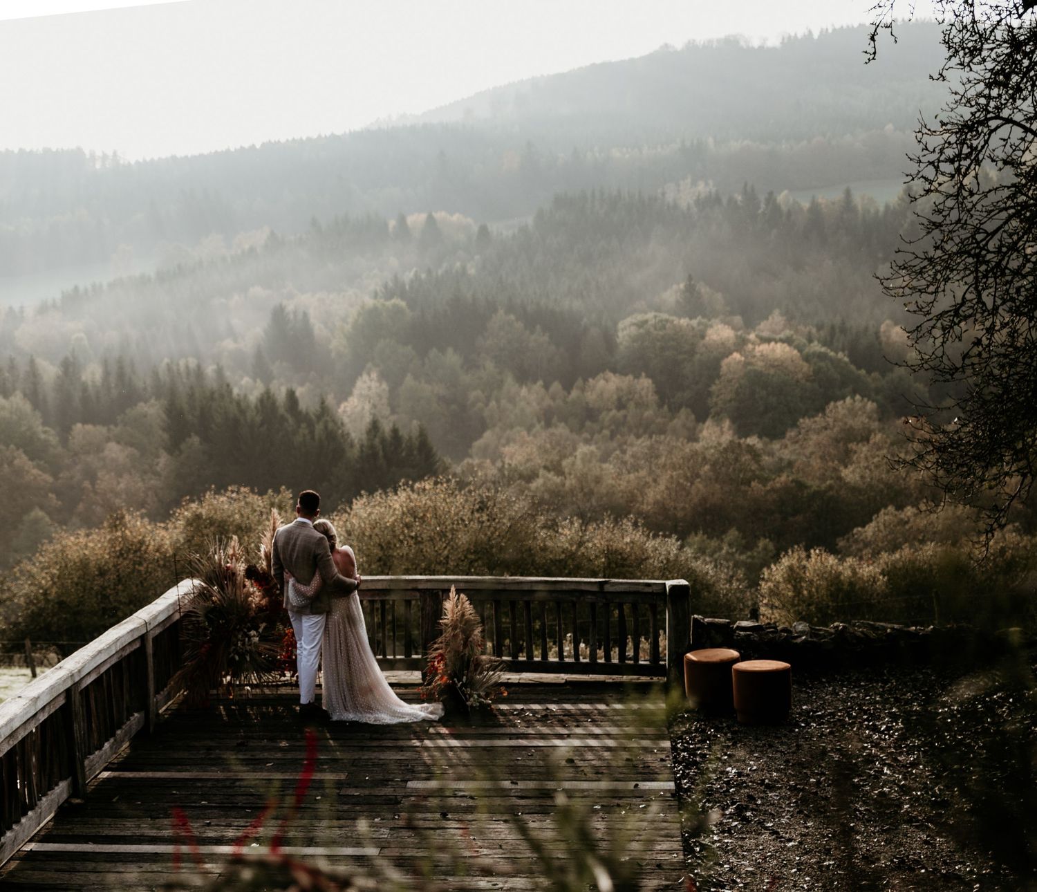 Couple standing on wooden deck overlooking misty mountain landscape at sunset.