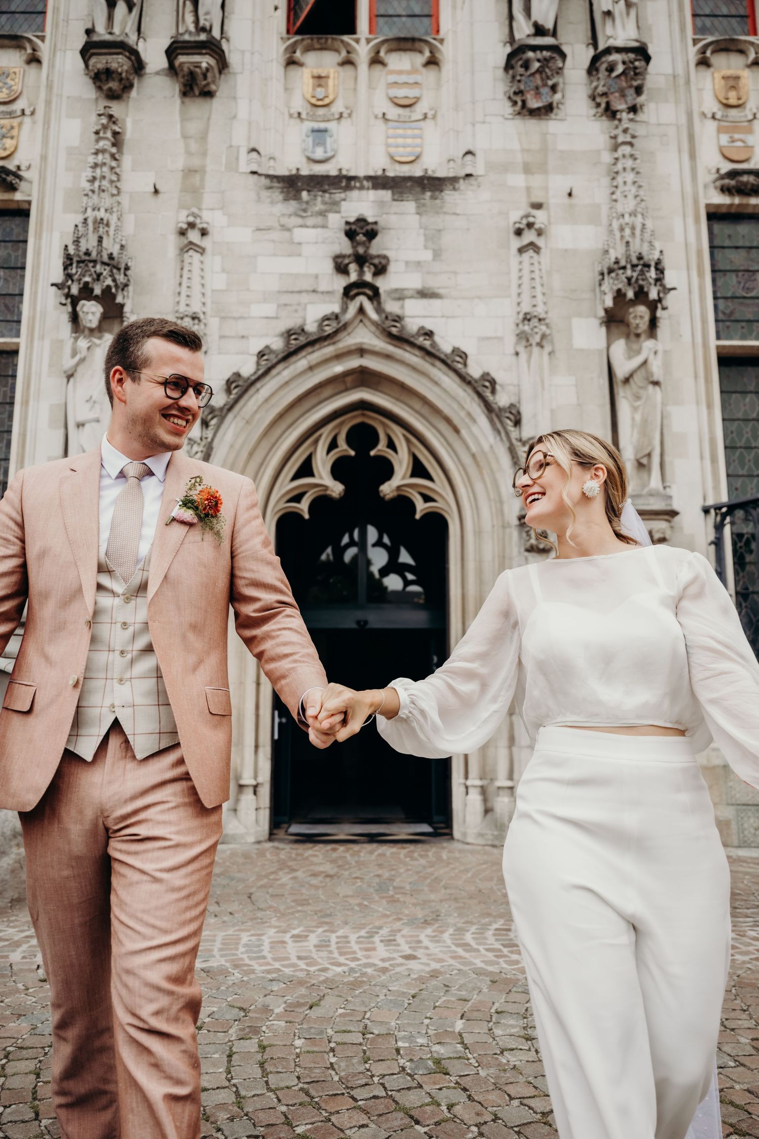 Modern couple in pink suit and white outfit dancing joyfully outside a gothic building.