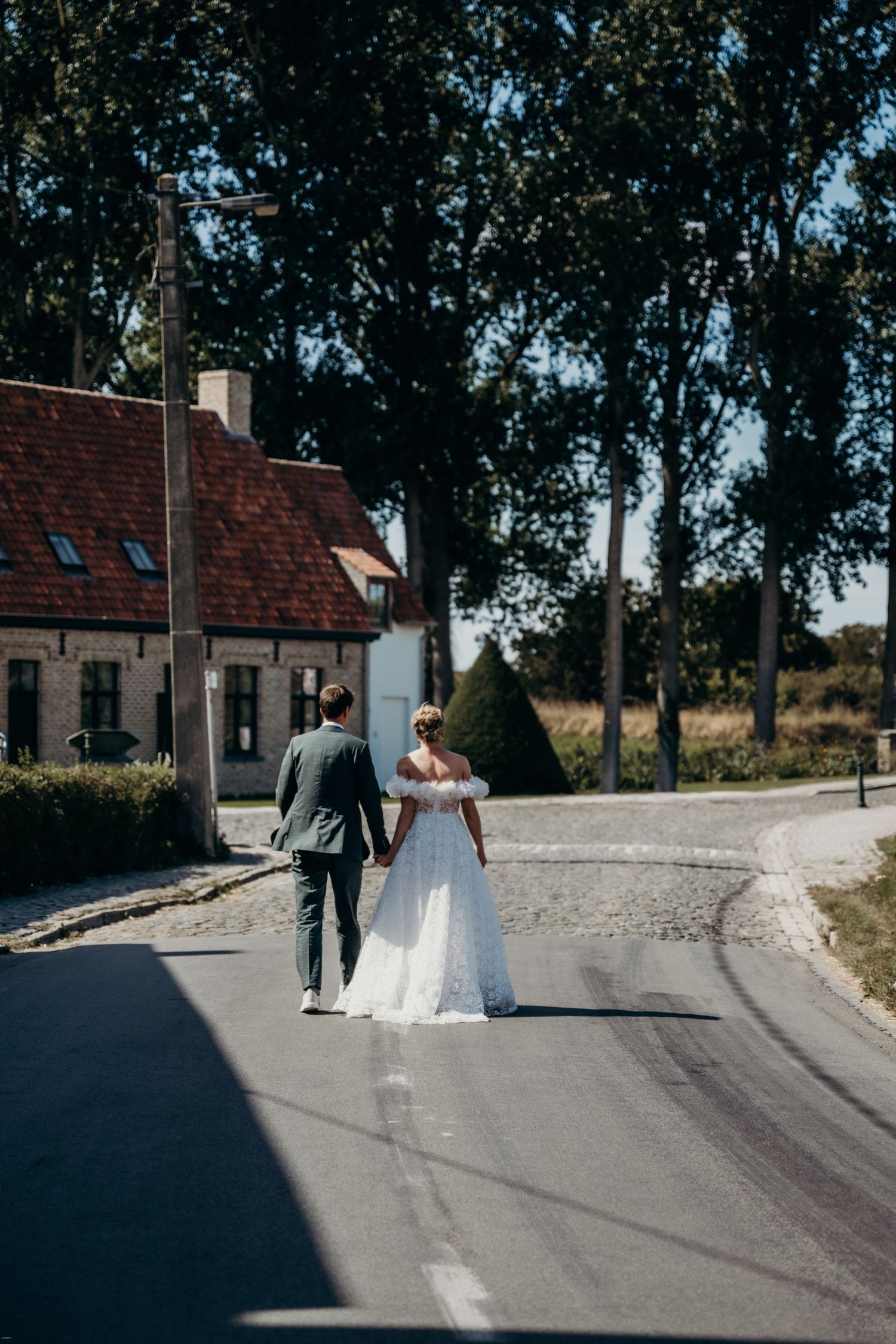 Couple in wedding attire walking down a country road towards a brick house.