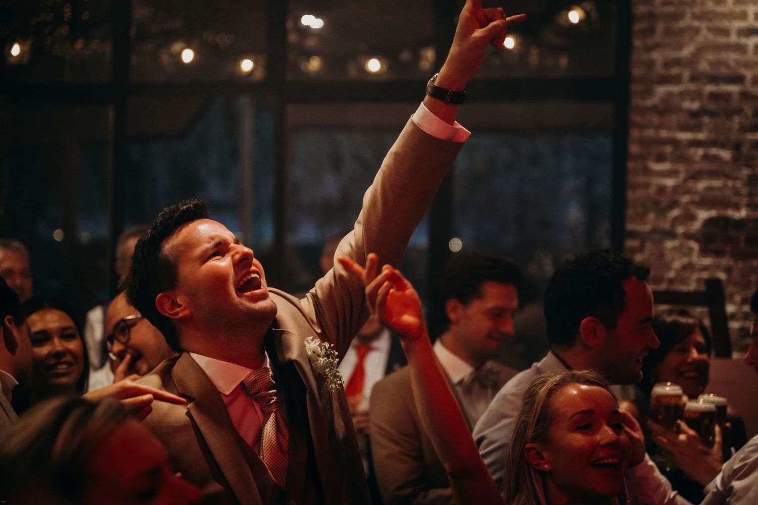 Wedding reception party crowd dancing under red stage lights in a brick-walled venue.