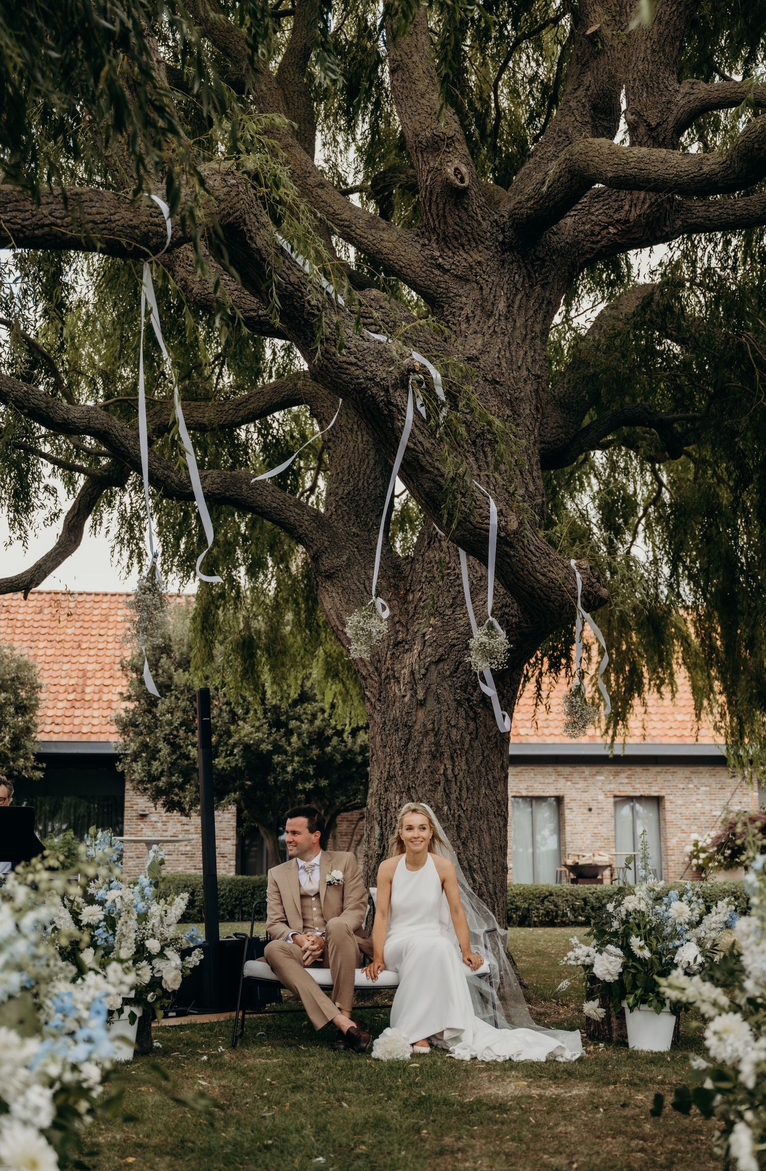 Wedding ceremony setup under a majestic willow tree with white floral arrangements.
