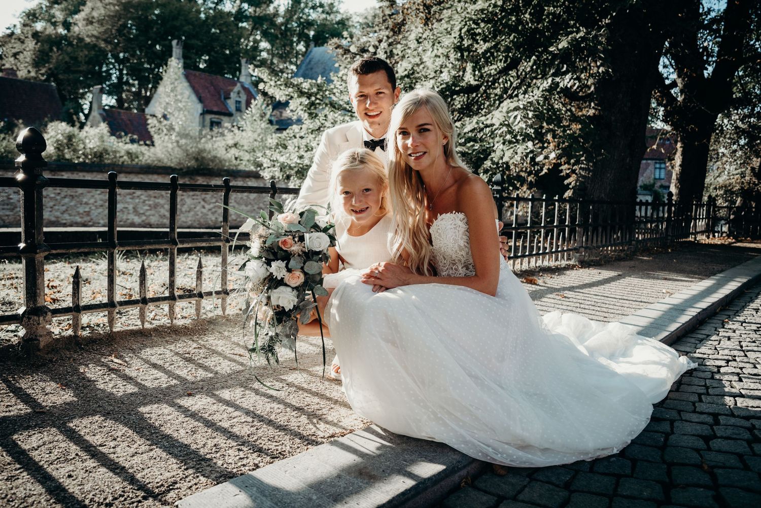A wedding couple poses with their child on a cobblestone path in a sunny outdoor setting.