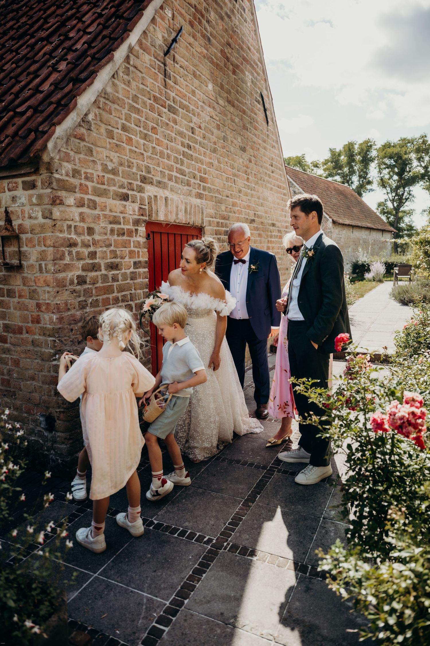 Wedding celebration with guests mingling outside brick barn surrounded by blooming garden flowers.