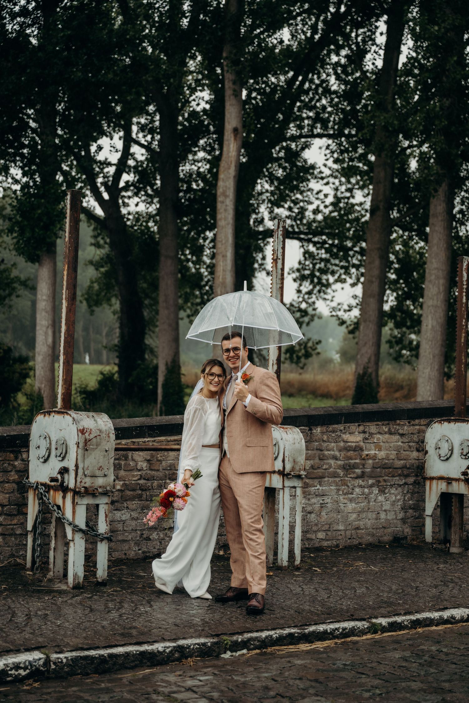 Wedding party poses under umbrellas against stone wall and tall trees in moody weather.