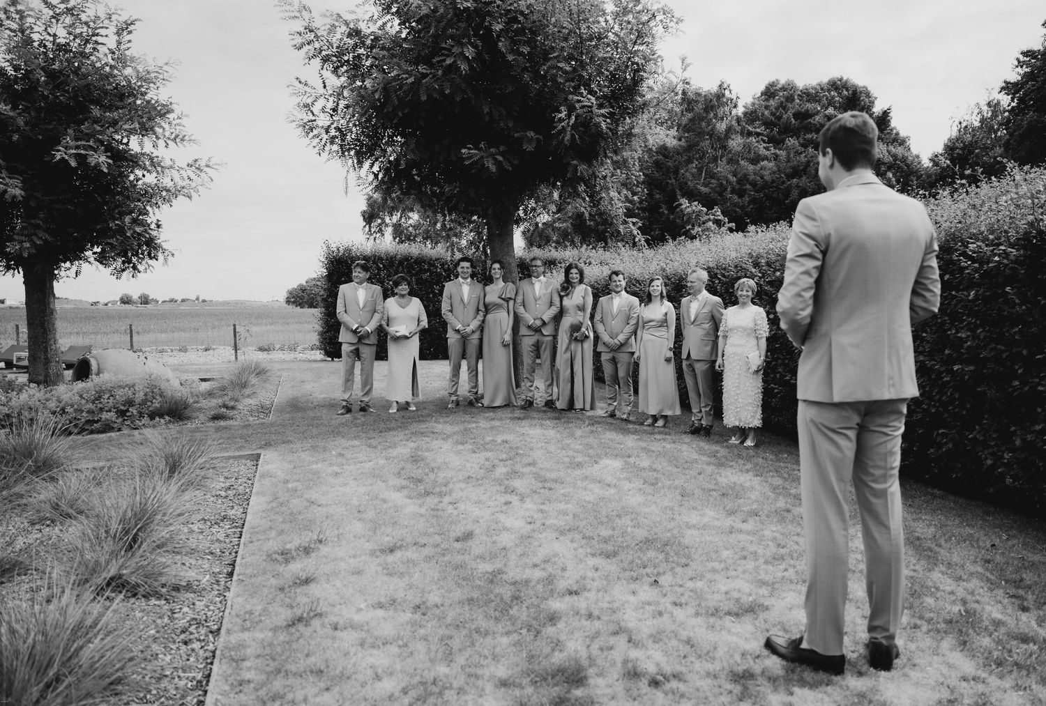 Wedding guests gathered outdoors on a grassy path lined with trees.