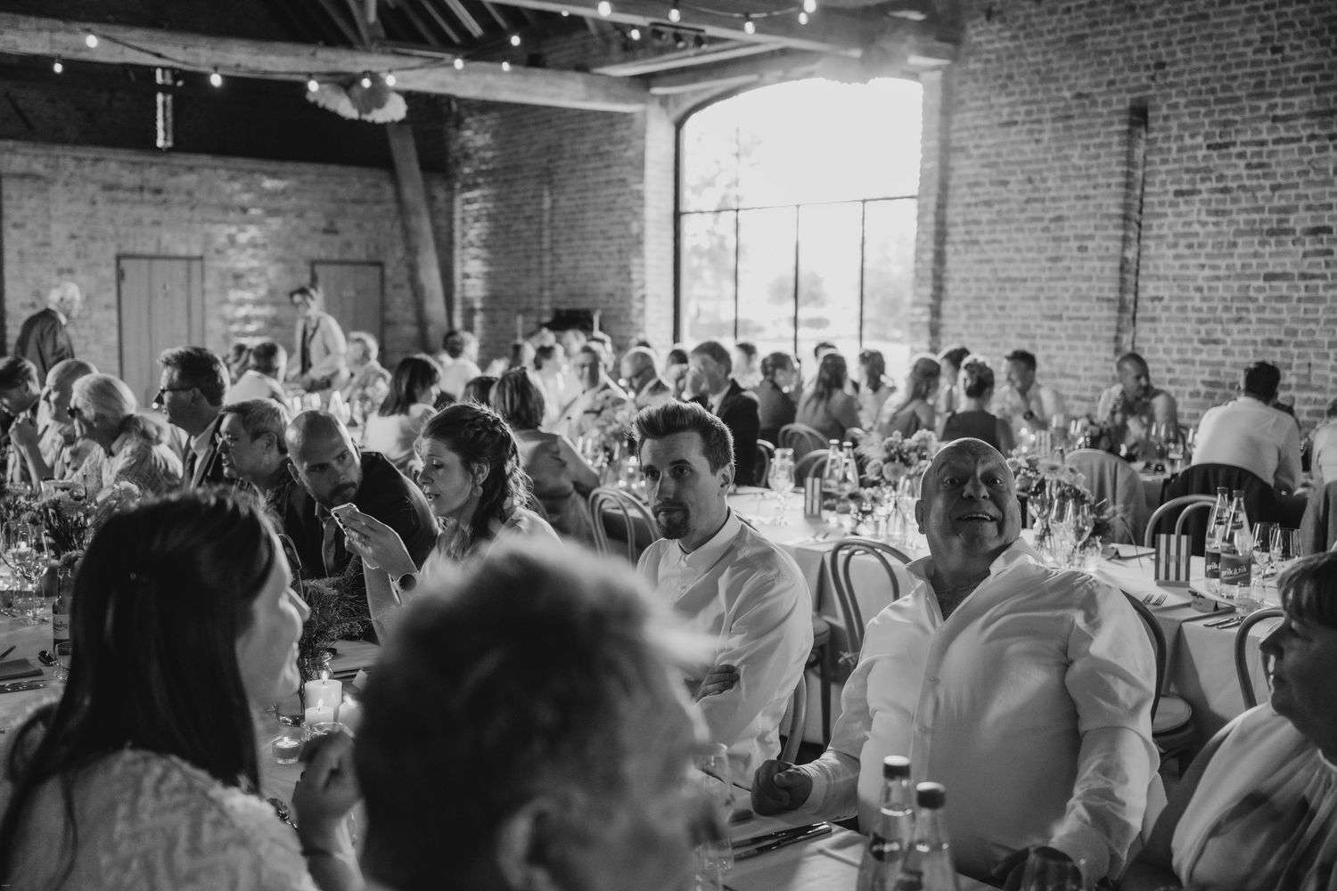 Black and white photo of wedding reception guests seated at tables in an industrial venue.