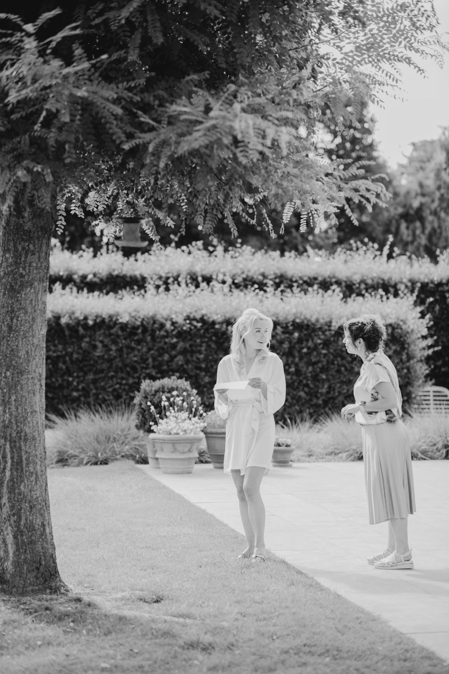 Wedding party members walking through a garden path carrying gifts and flowers in black and white.