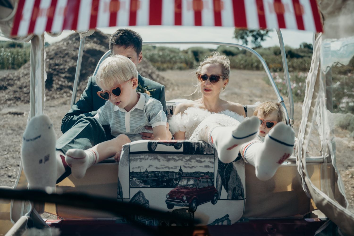 Wedding party relaxes in a vintage-style vehicle under a striped awning on a sunny day.