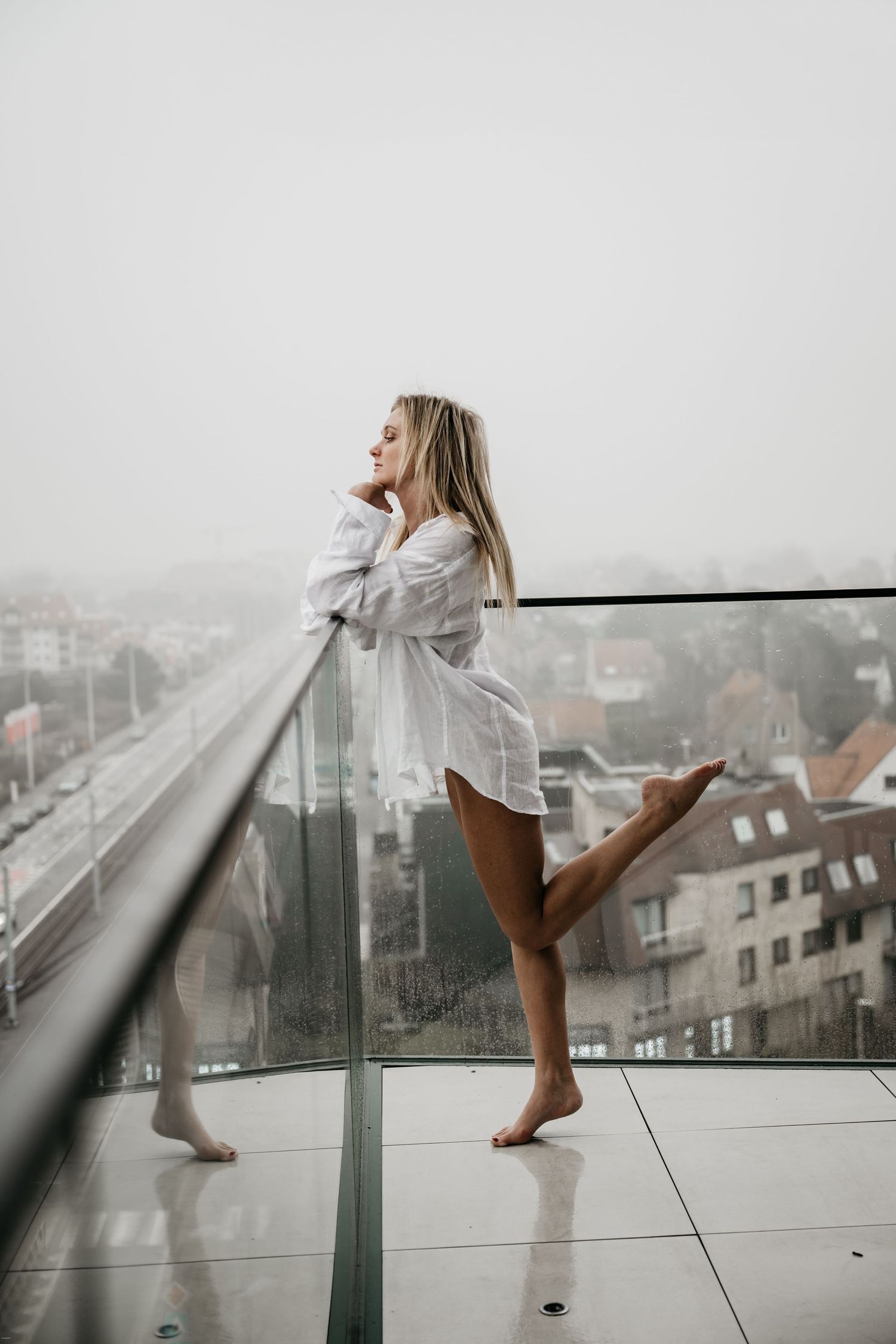 Woman in oversized white shirt dances on balcony overlooking foggy cityscape.