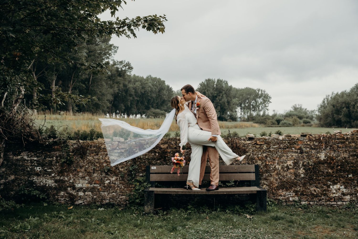 Couple sits on rustic stone wall during outdoor wedding photoshoot.