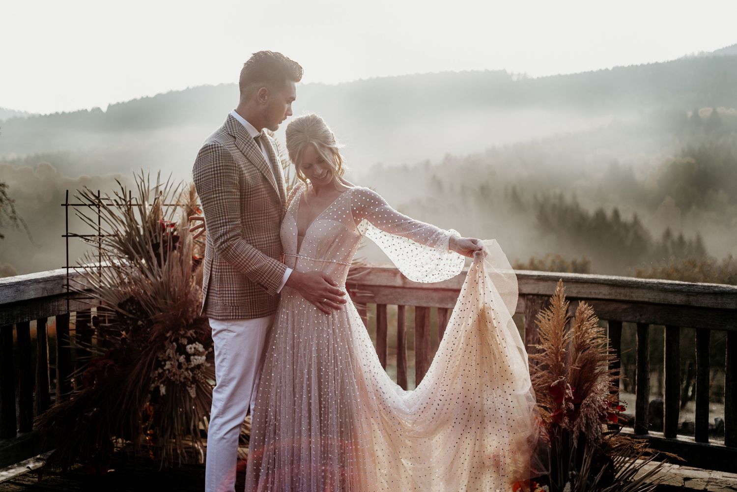 Wedding couple poses against mountain backdrop on rustic wooden deck with autumn decorations.