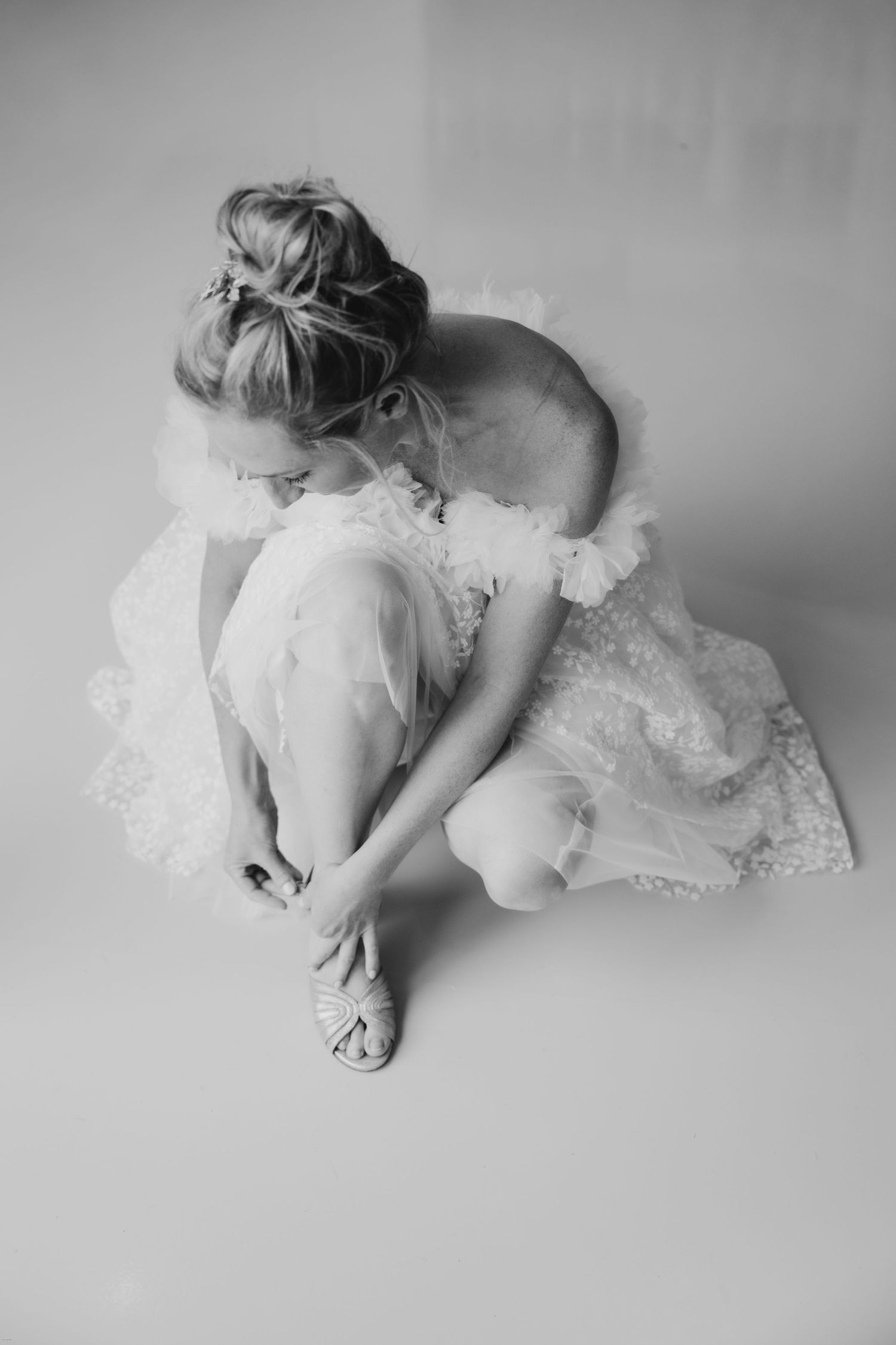 A bride in a white lace dress adjusts her shoe in an artistic black and white portrait.