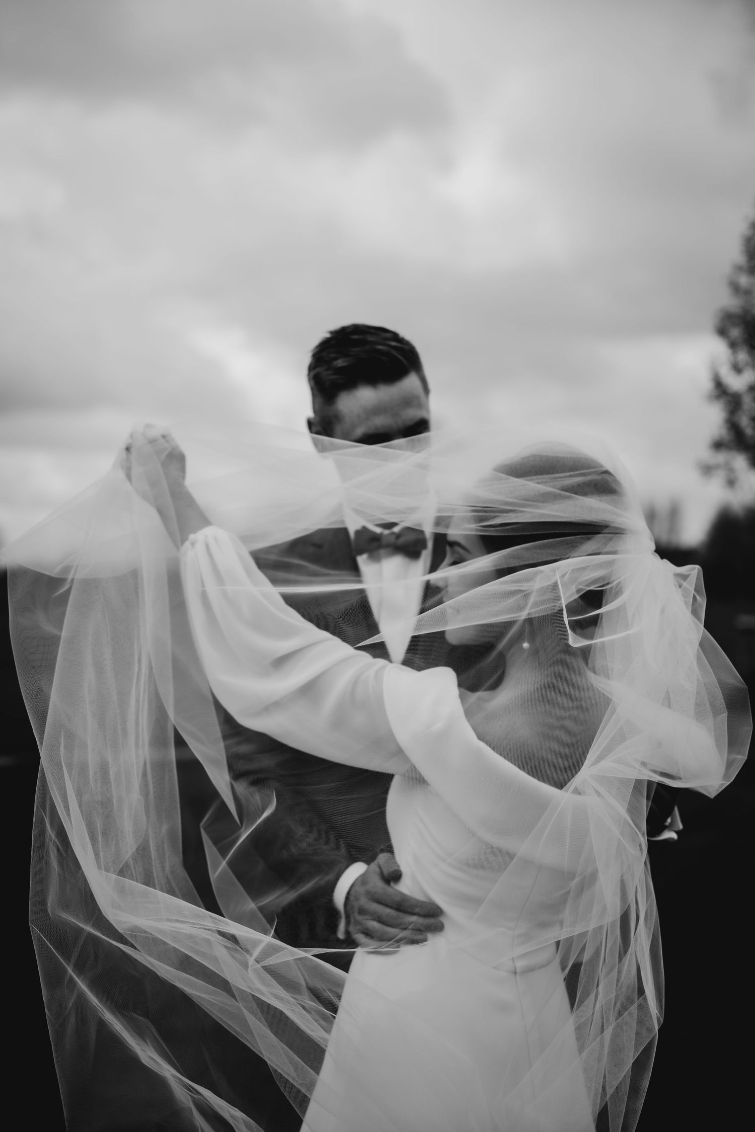 A flowing white wedding veil blows dramatically in the wind against a cloudy sky.