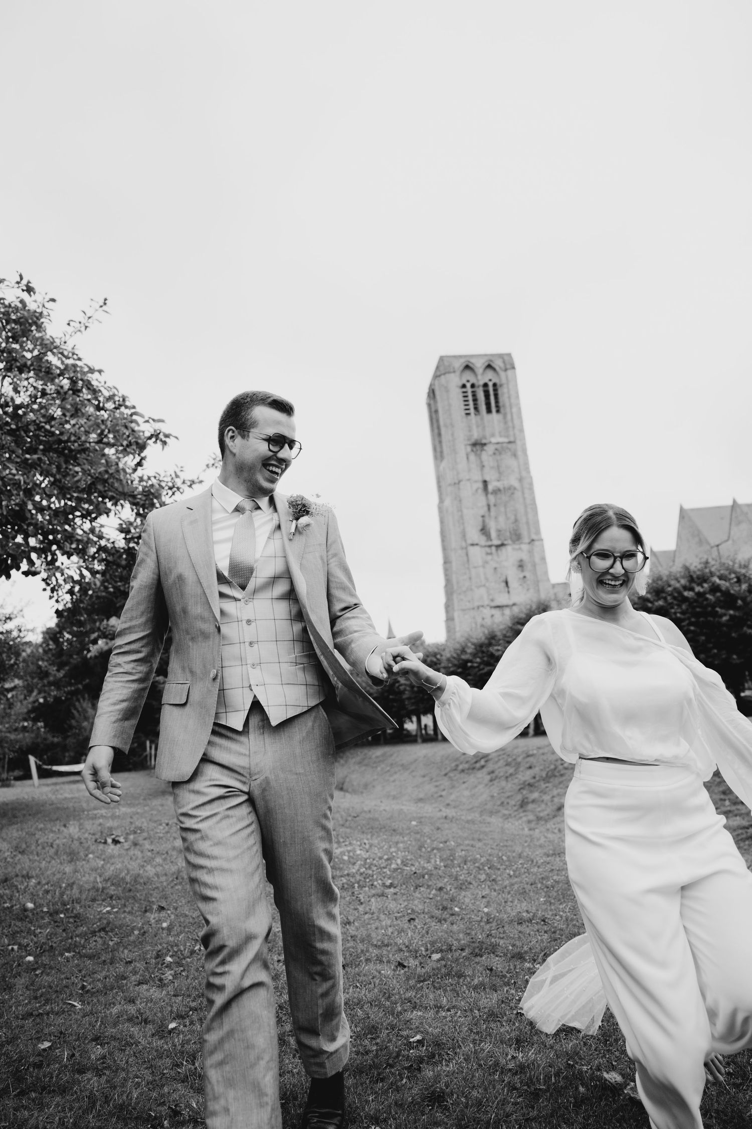 Wedding couple running and playing together on grass with a church tower in the background.