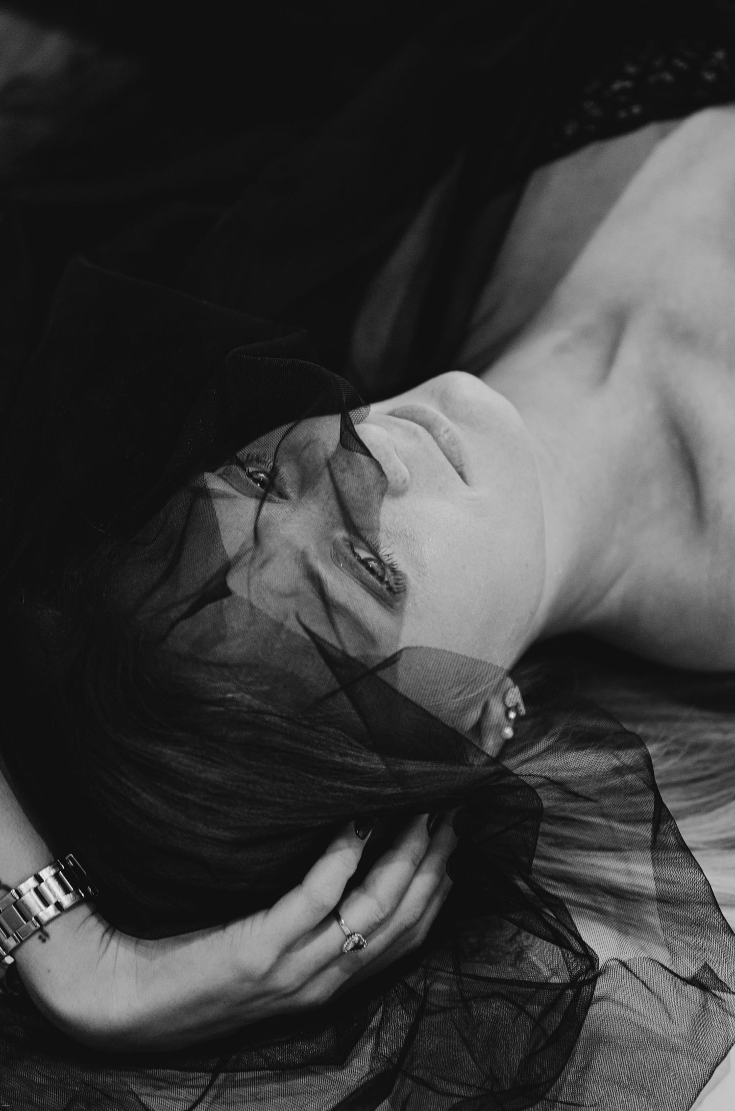 Black and white artistic portrait showing person lying down with dark hair and sheer fabric against dark background.