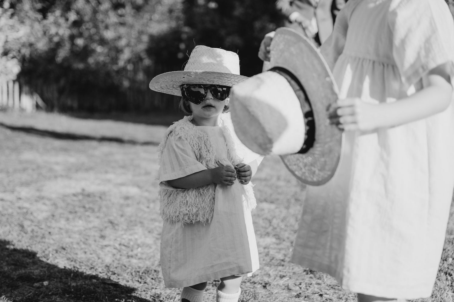 A small child in sunglasses and a hat stands near a drum at an outdoor event.