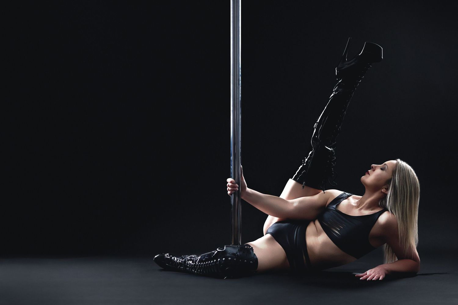 Woman in black two-piece lingerie and thigh-high boots while leaning back on a dance pole during a South End, Charlotte, boudoir session.