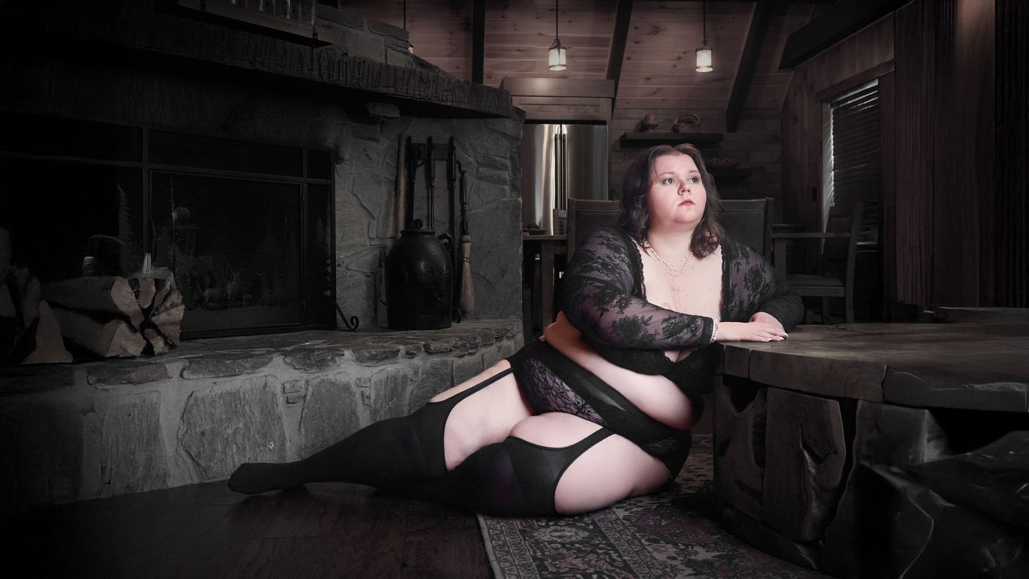 Plus-size boudoir in Charlotte capturing an image of a woman lying against a table by a fireplace in a cabin.