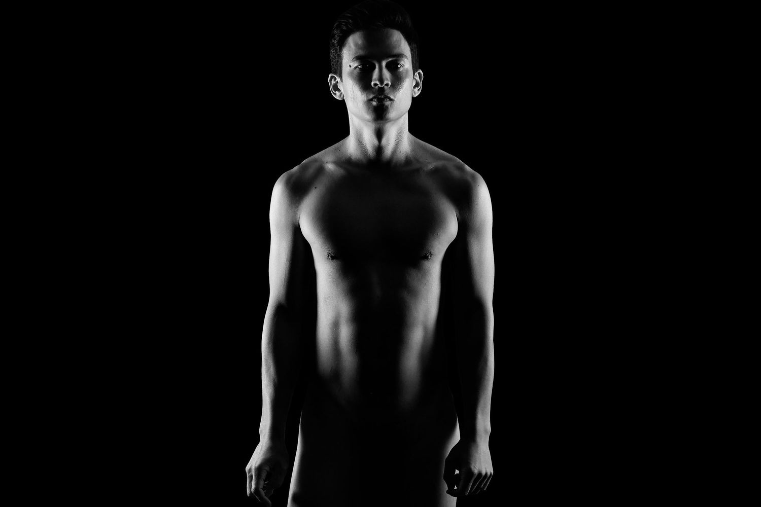 A high-end male boudoir photo of a man posing nude with detailed lighting and a dark background.