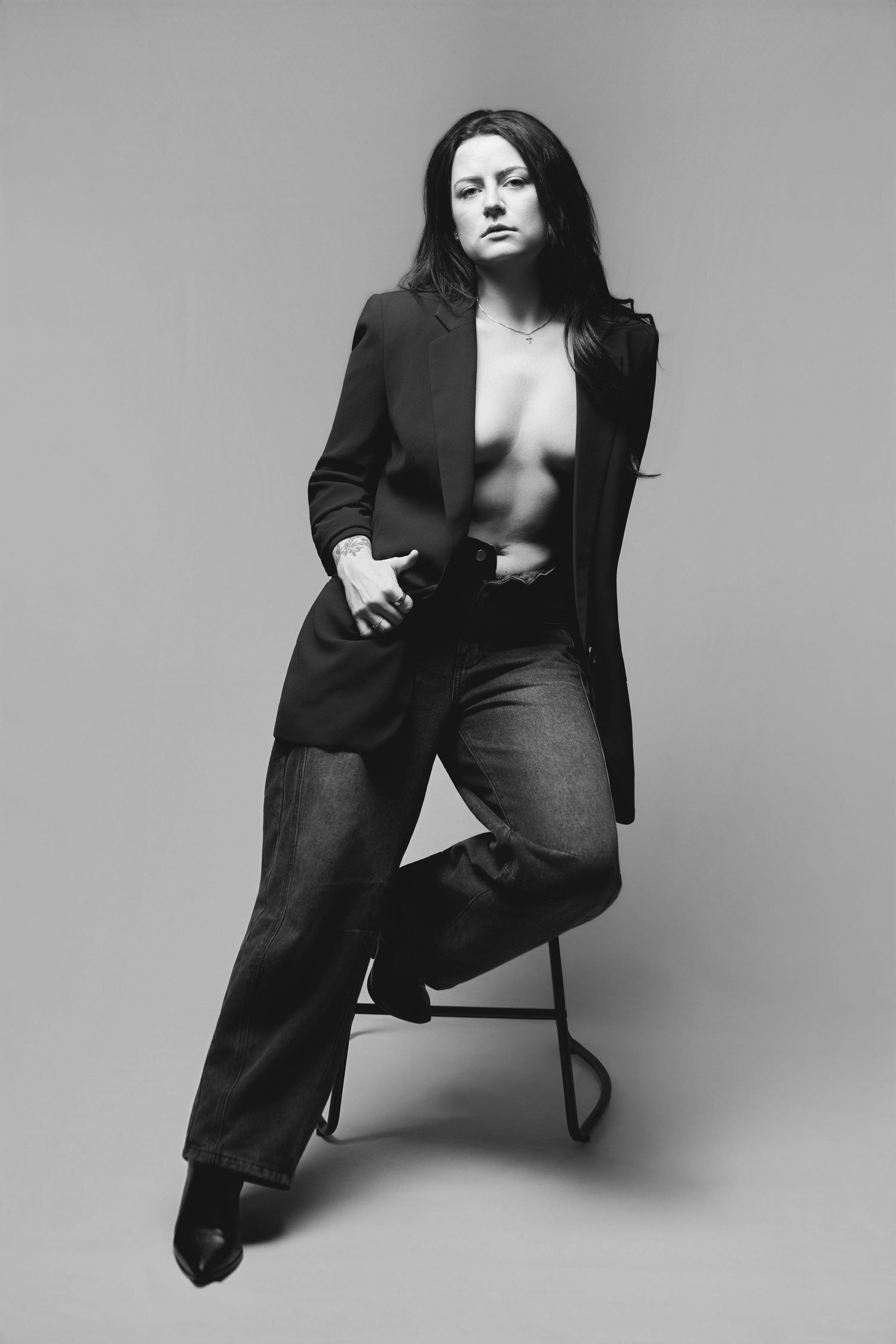 Black and white artistic portrait of a person in a blazer and jeans posing on a stool against a plain backdrop.
