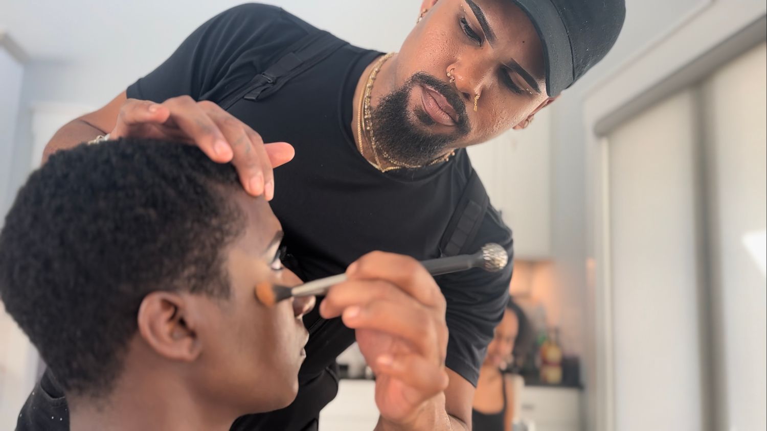 A professional makeup artist, wearing black attire and a cap, applies cosmetics to a client in a bright studio setting.
