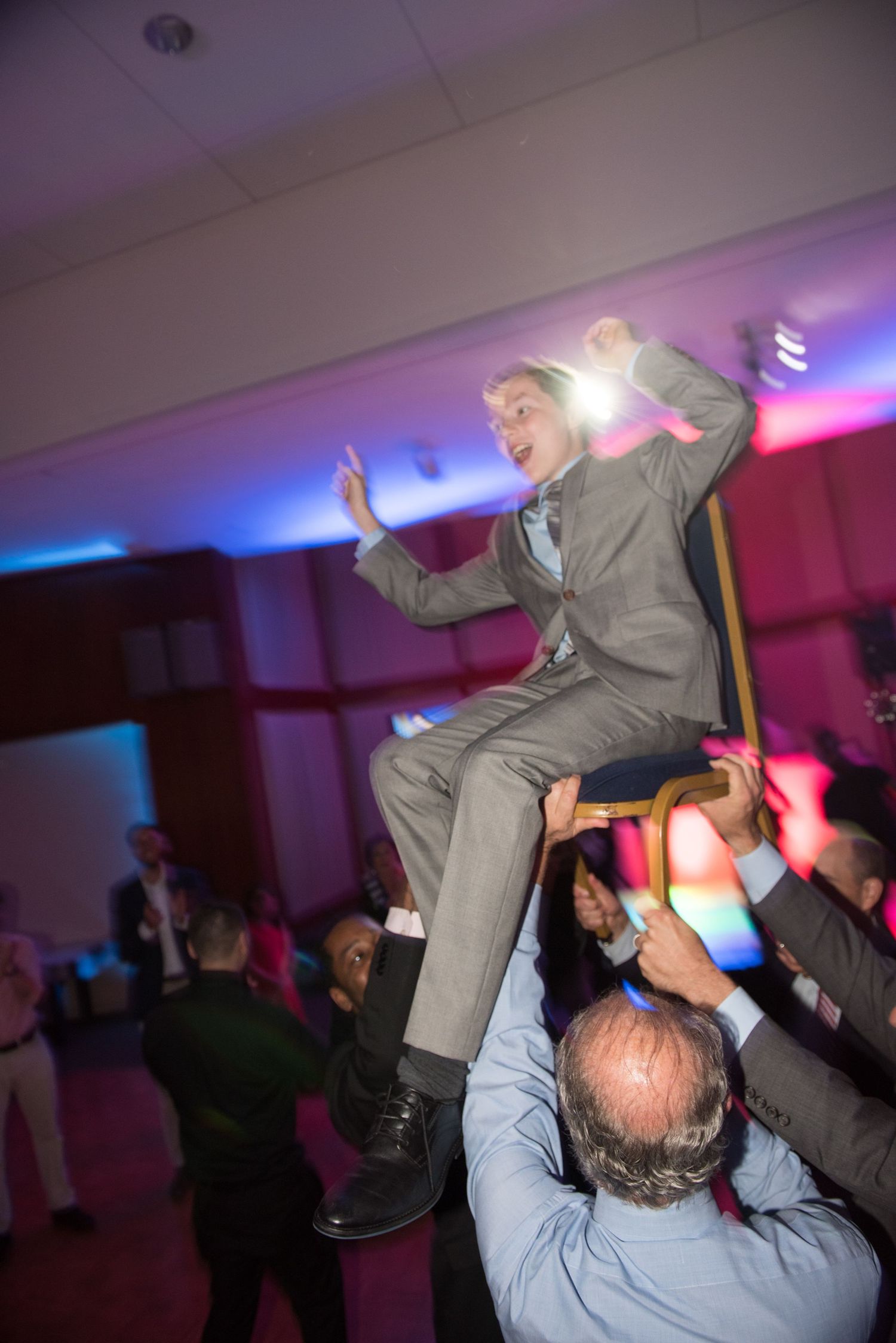 Energetic dance party moment with someone being lifted in the air on a purple-lit dance floor.