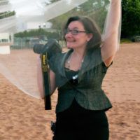 A joyful person in a green vest raises their arms in celebration on a sandy beach.