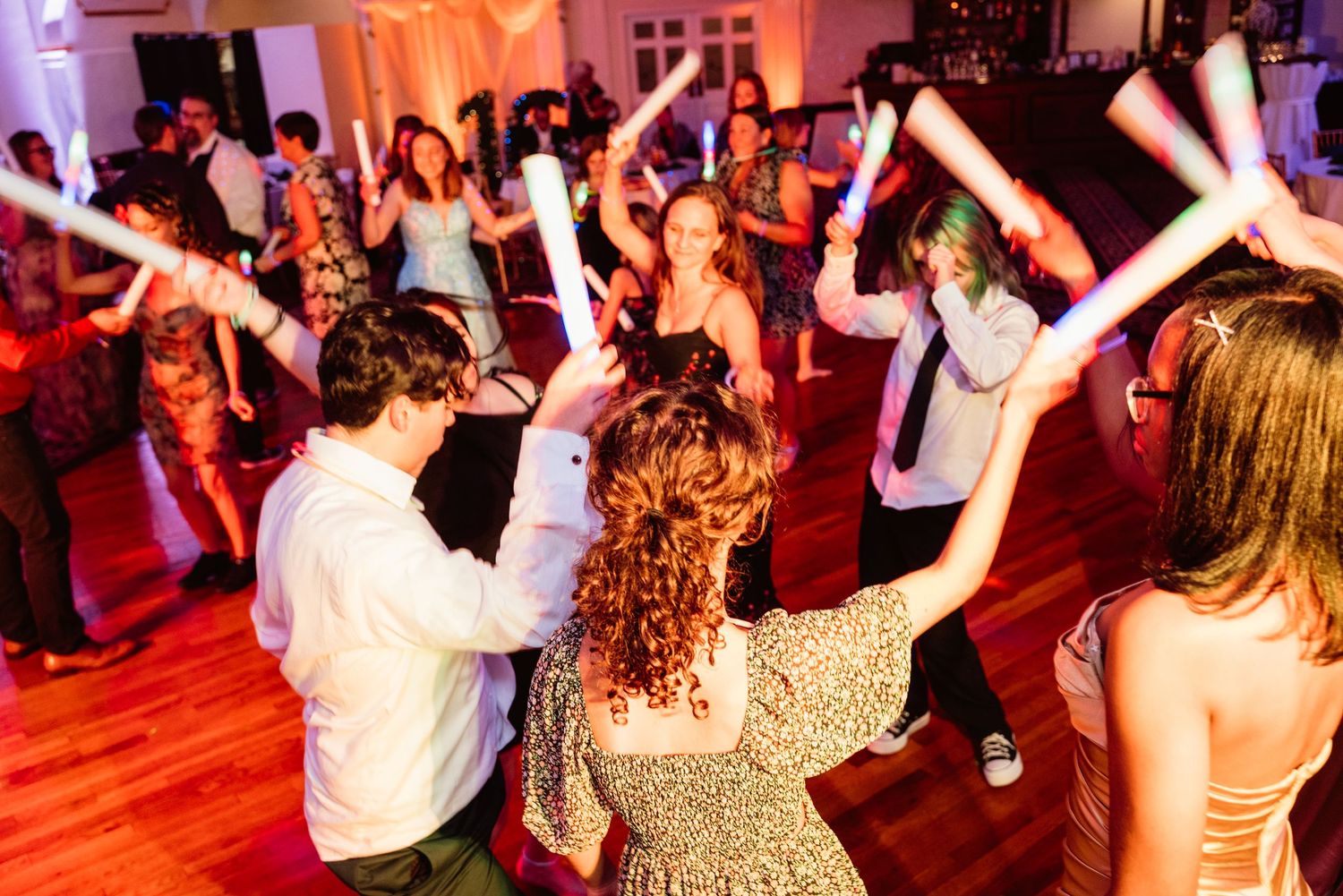 Crowd dancing with raised arms at a wedding reception with pink and purple lighting.