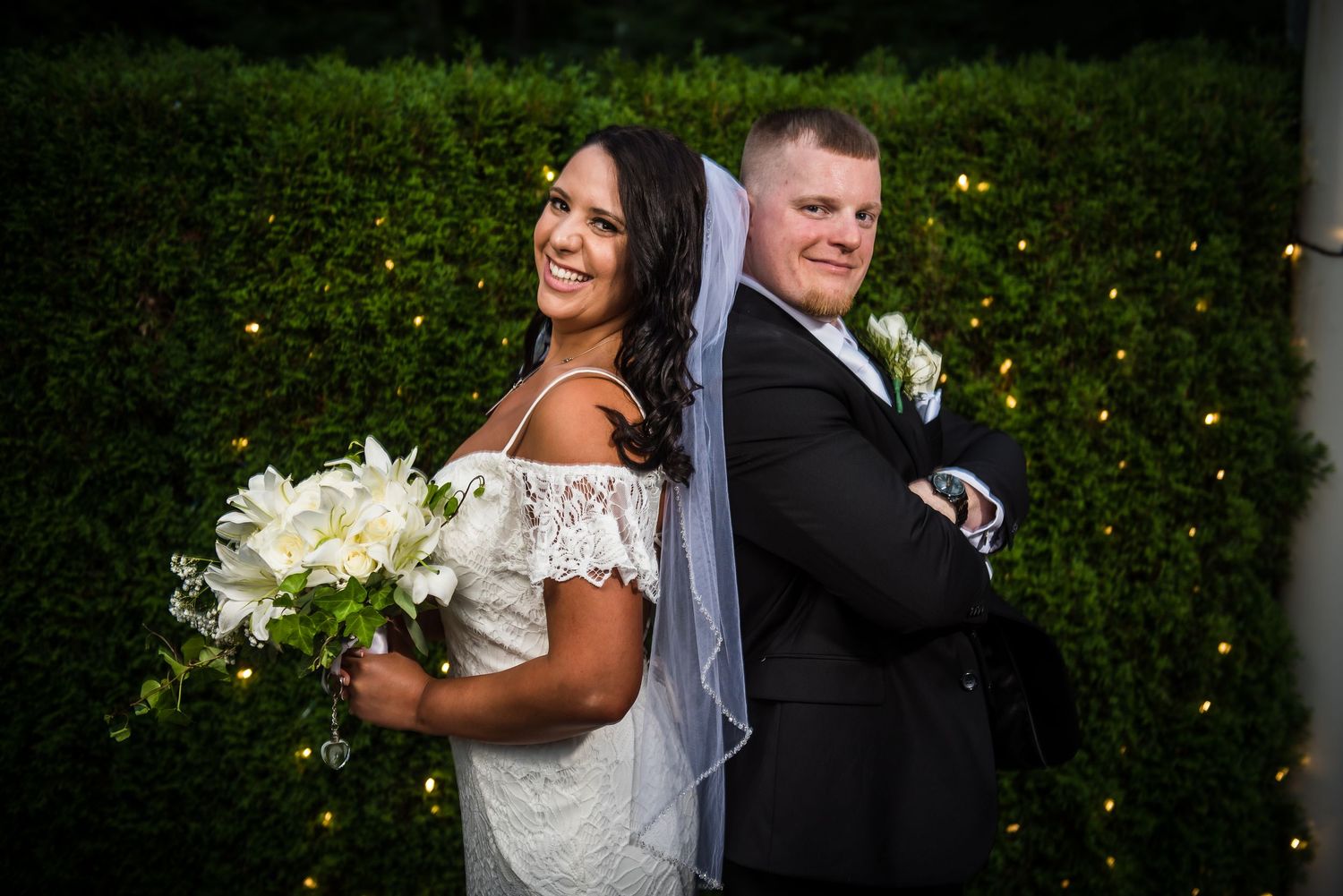 Wedding party members pose together outdoors at dusk with decorative lights in the background.