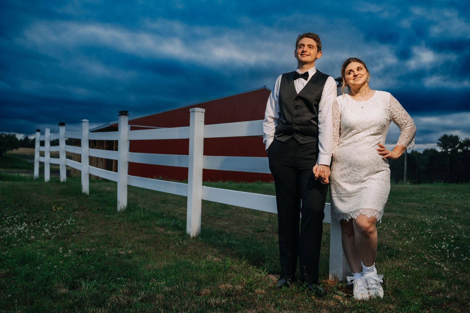 Wedding couple posing against dramatic blue sky and white fence at sunset.