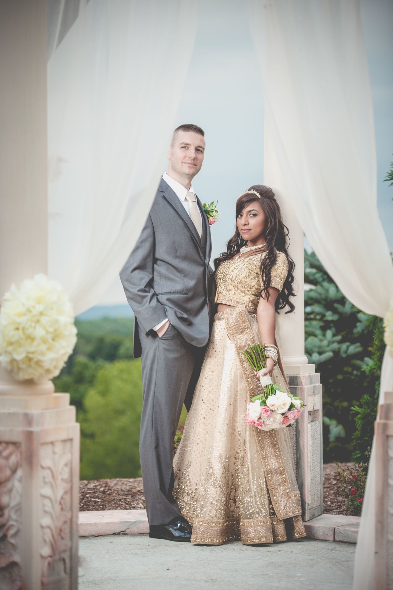 Wedding couple poses outdoors with bride in gold dress and groom in grey suit near white curtains.