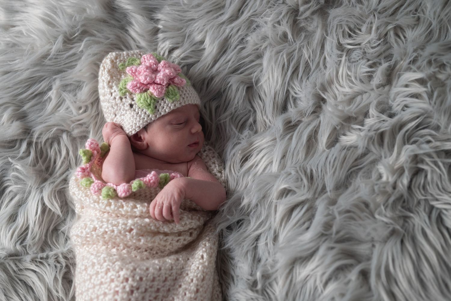 Newborn baby wearing a crocheted dinosaur outfit lying on soft grey fur blanket.