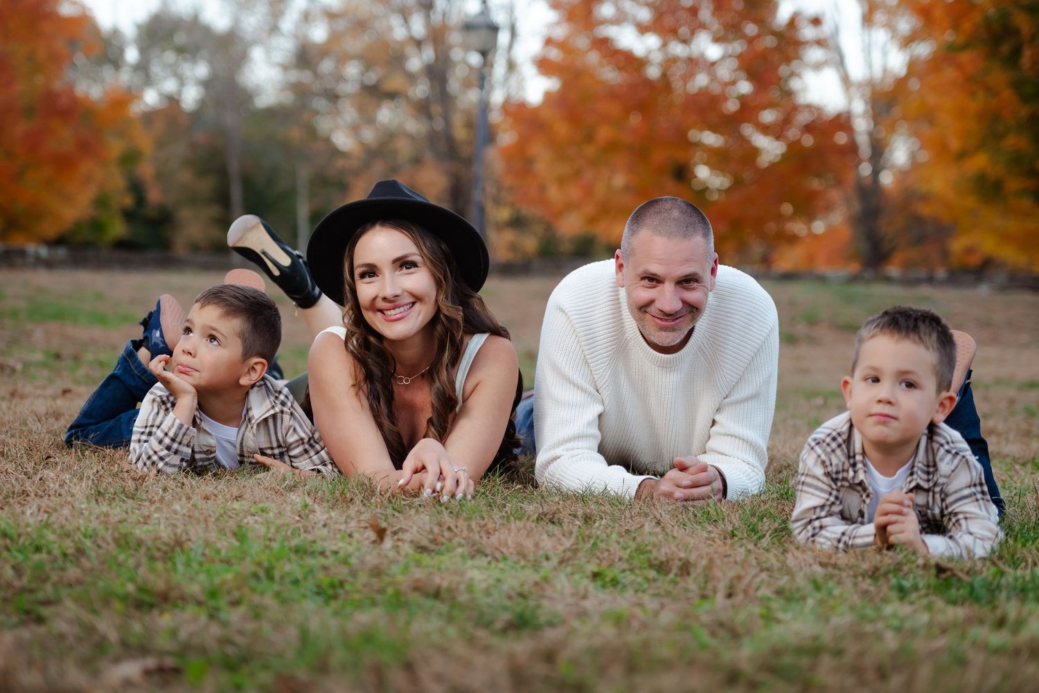 Family lying on grass during autumn with colorful fall foliage in background.