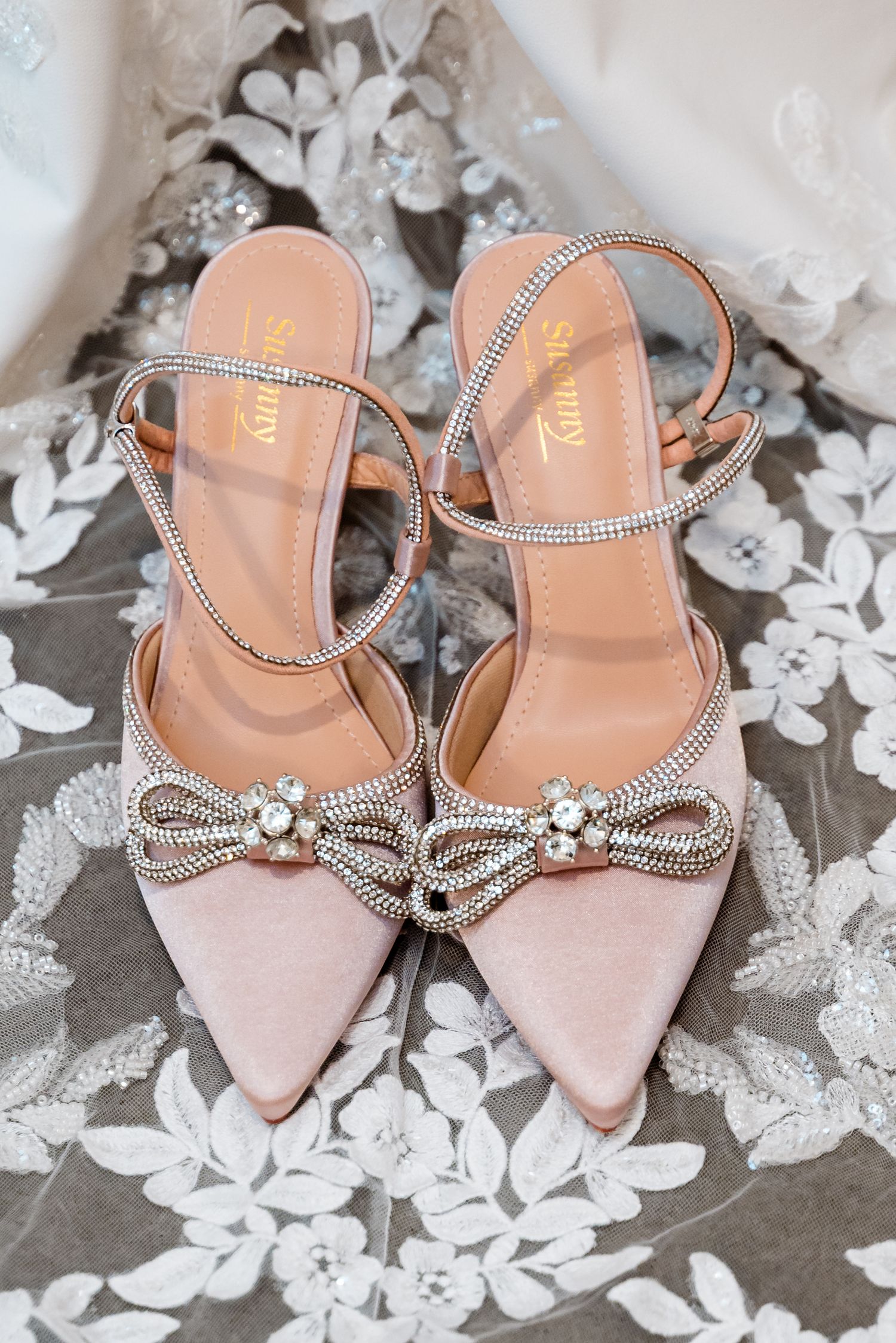 Blush pink bridal shoes with crystal embellishments displayed on lace fabric.
