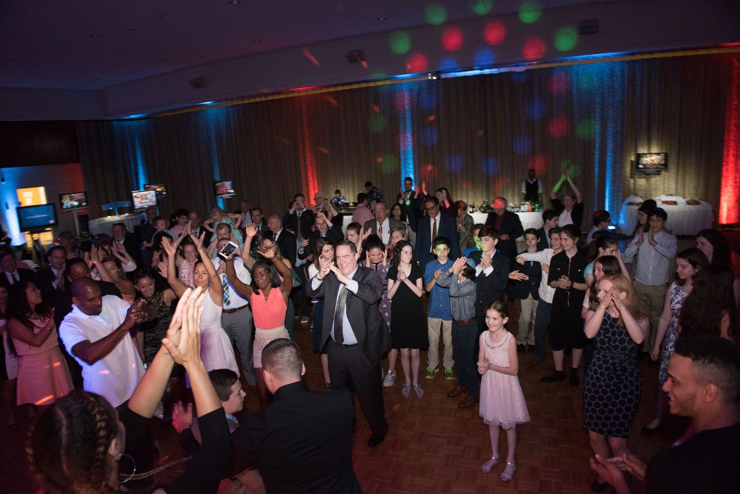 Large group dancing and celebrating under colorful party lights in a reception hall.