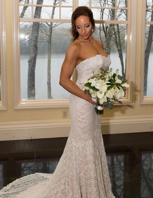 Bride in elegant white lace gown holds a classic white bouquet by sunlit windows.