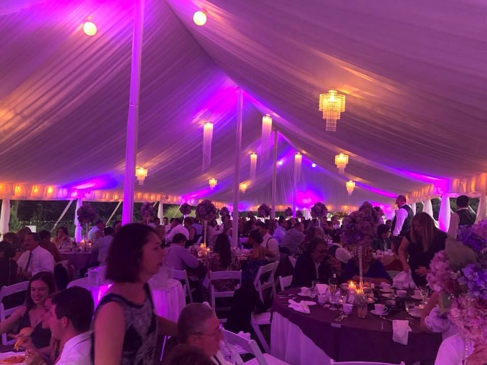 Elegant wedding reception venue with purple uplighting and chandeliers illuminating tables of guests in a large tent.