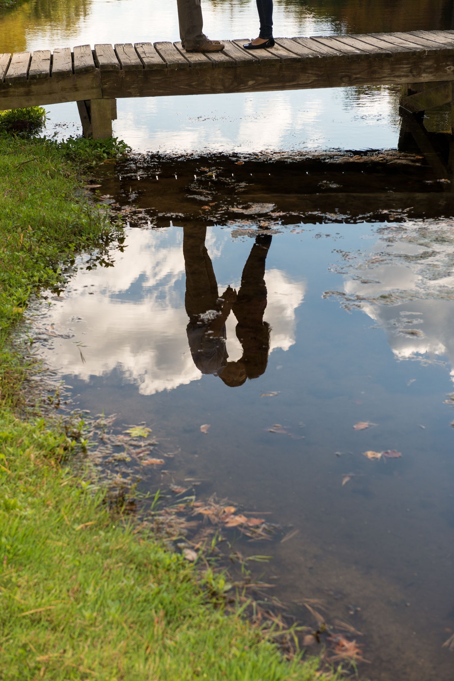 A reflection in water shows two people standing on a wooden footbridge.