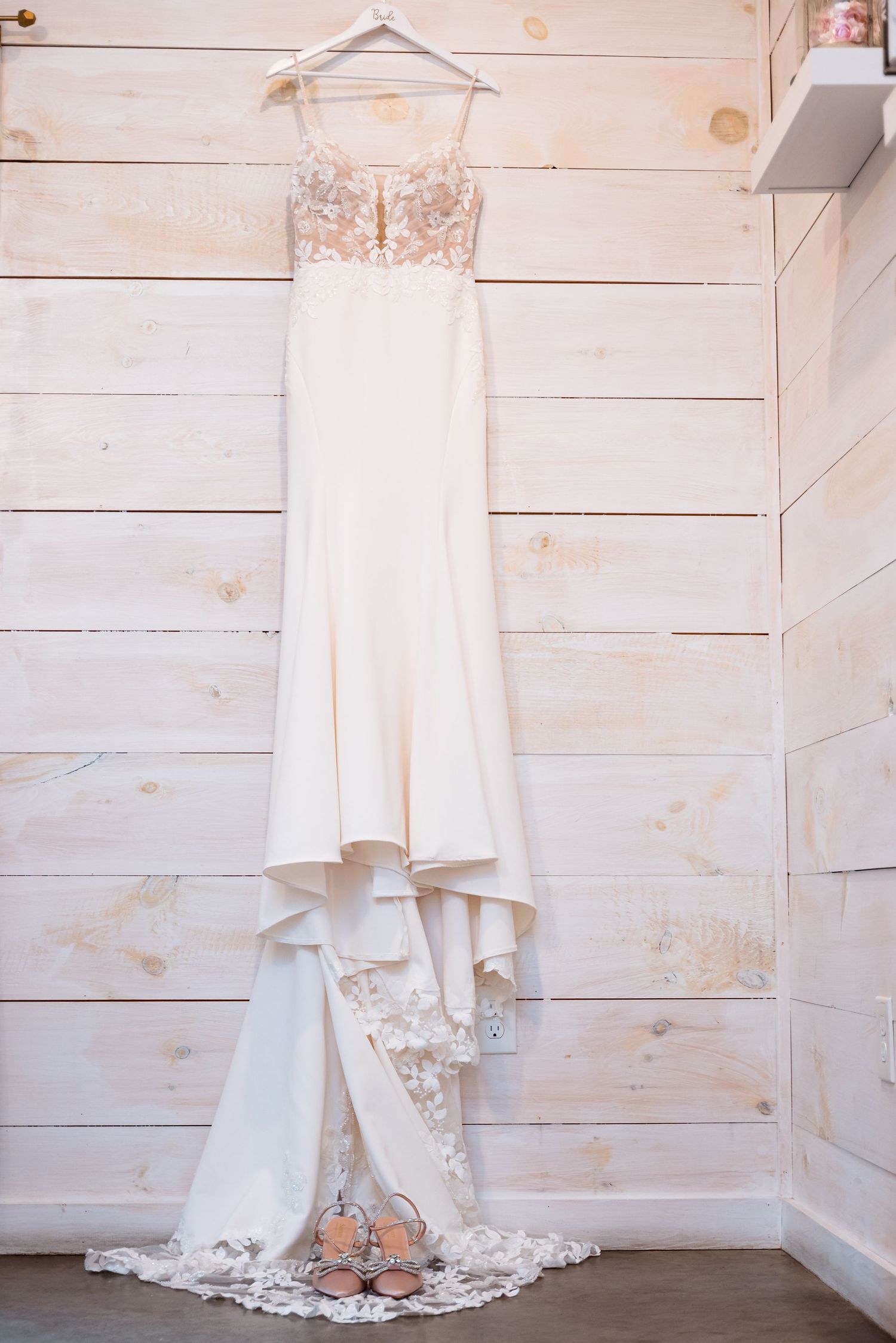 White lace wedding dress hanging against rustic wooden wall background.