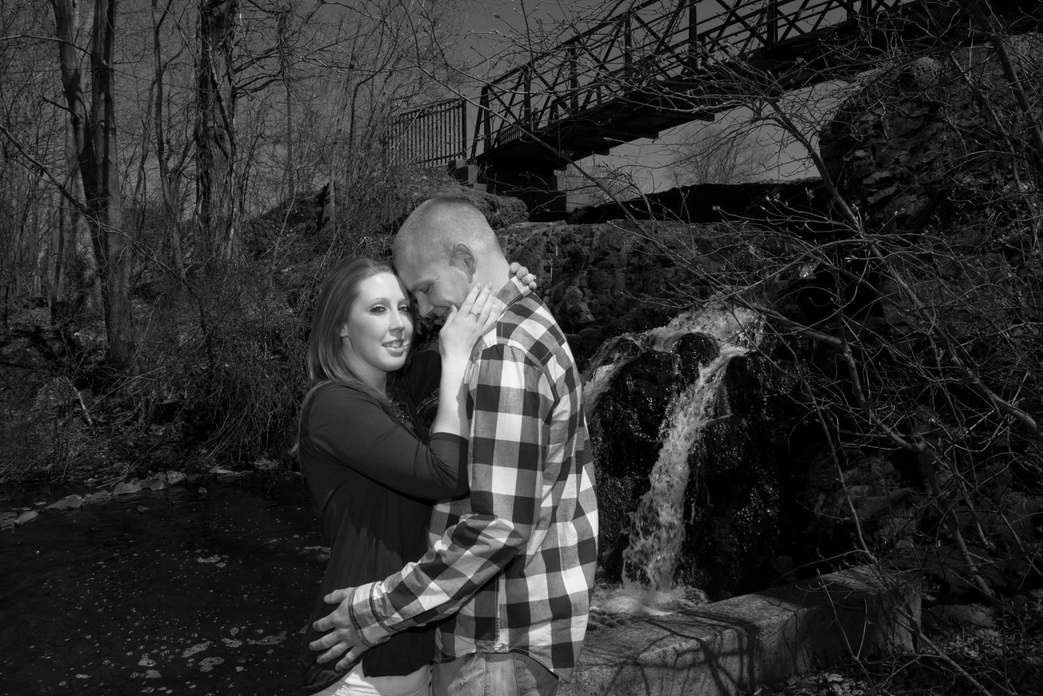 A couple embracing near a rustic bridge in a moody black and white photograph.