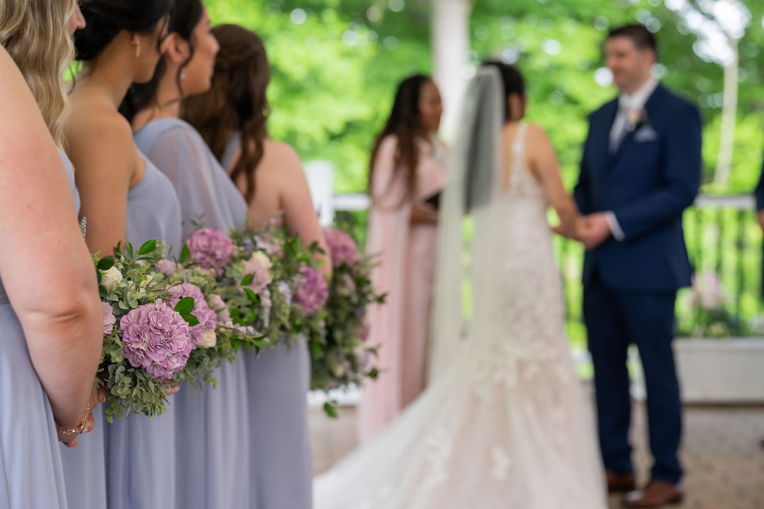 Bridesmaids in lavender dresses hold pink bouquets during an outdoor wedding ceremony.