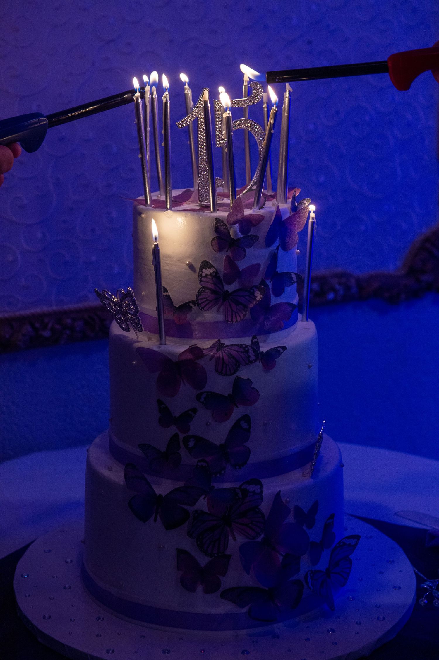 A white tiered cake with purple butterfly decorations glows with lit candles.