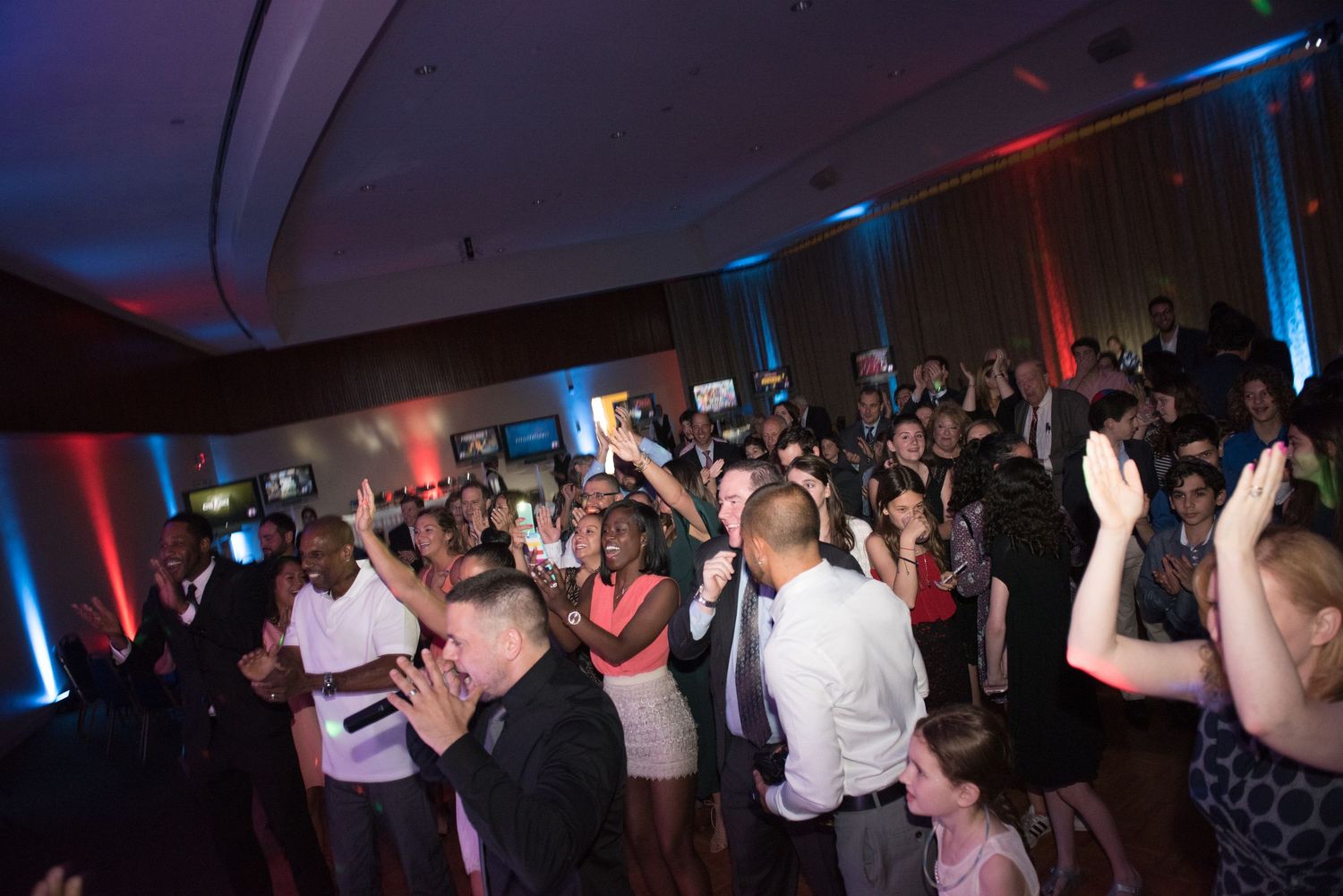 Crowd of people dancing with raised hands under blue and red lighting at an event.