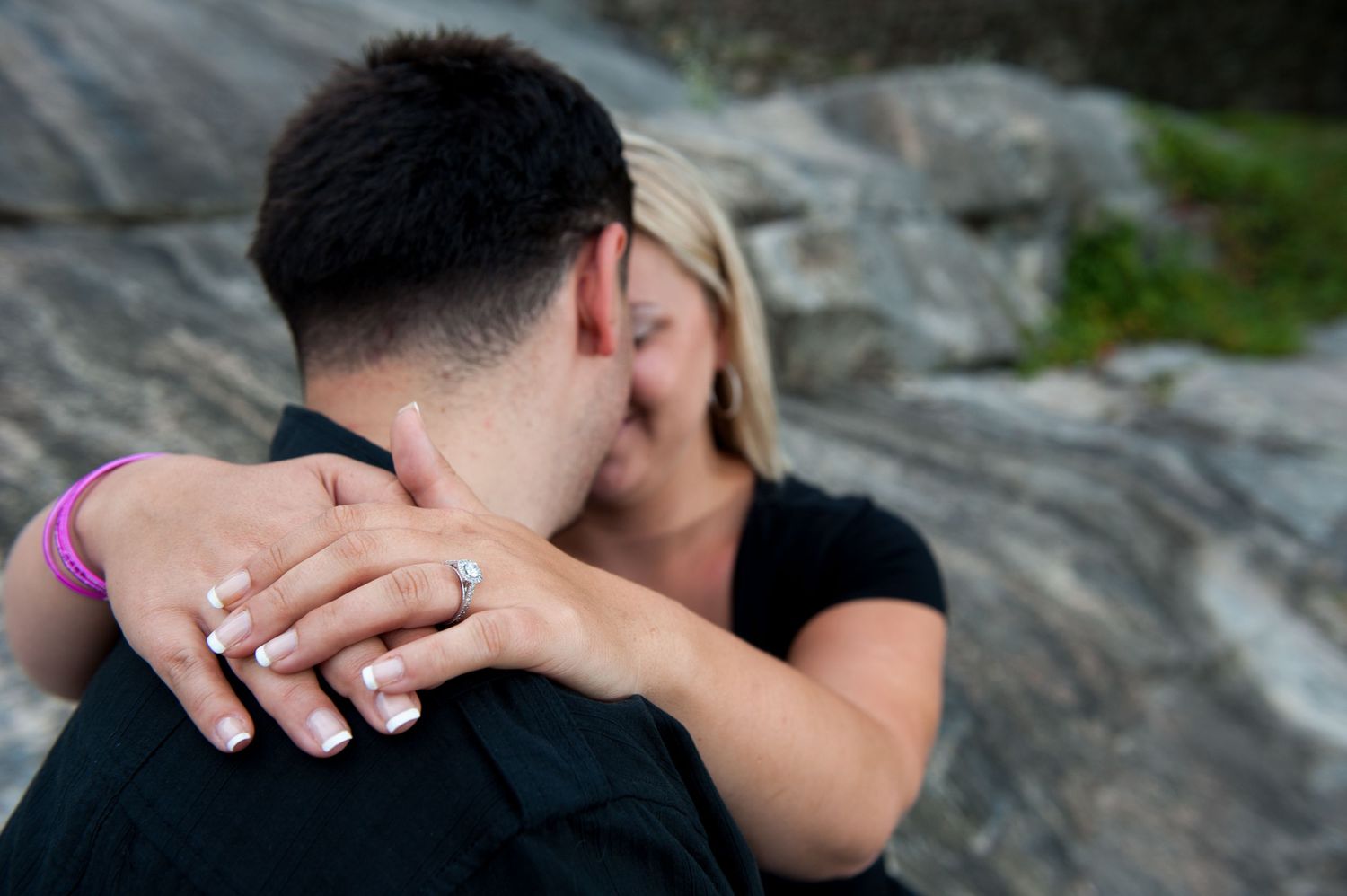 Close-up of an engagement ring while a couple shares an intimate moment on rocks.