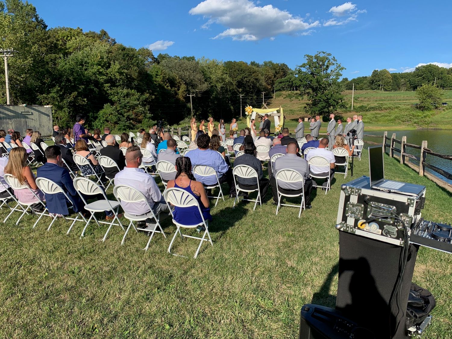 Outdoor wedding ceremony setup with white chairs on a grassy hill under blue summer skies.
