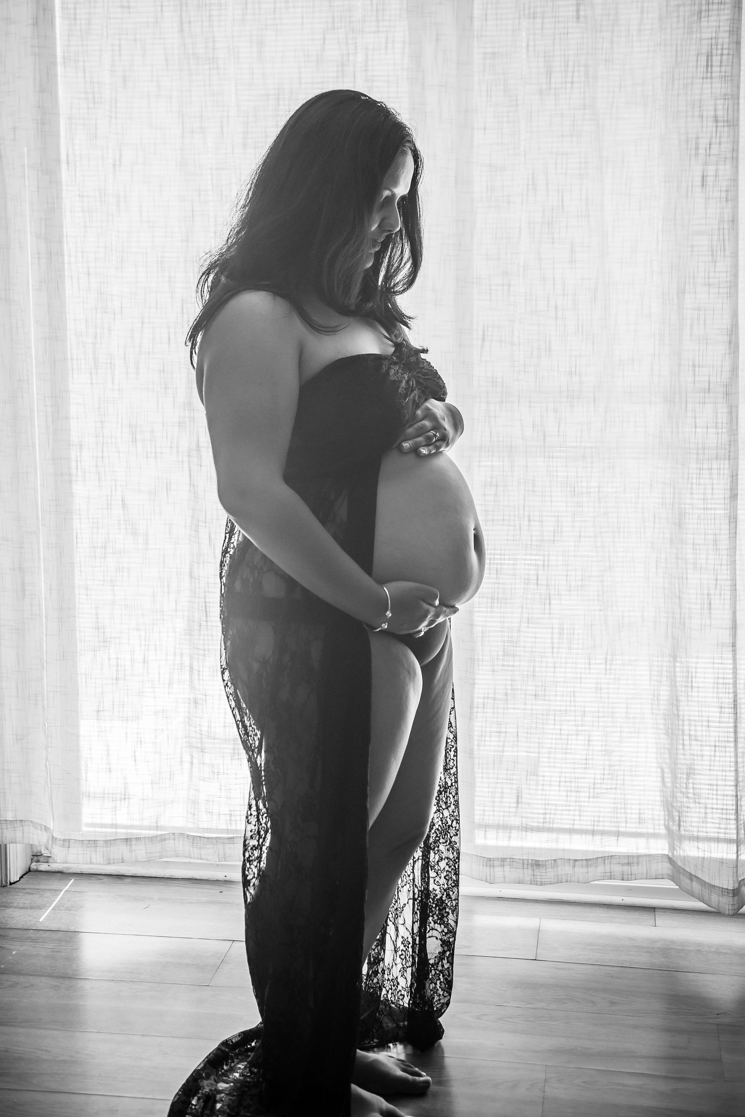 Silhouette of pregnant woman in flowing dress against white curtained window.