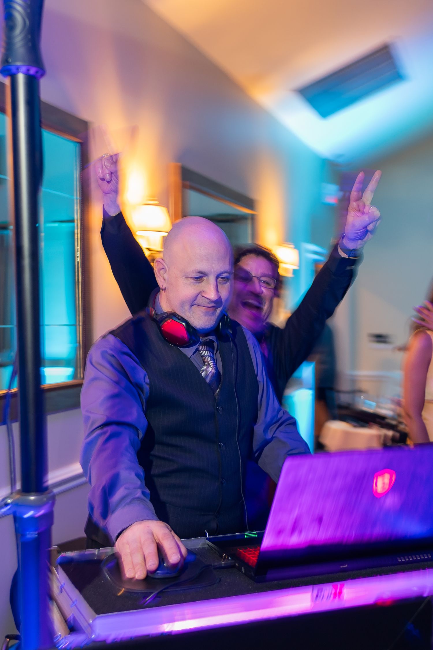 DJ raises hands while performing behind equipment with colorful venue lighting.