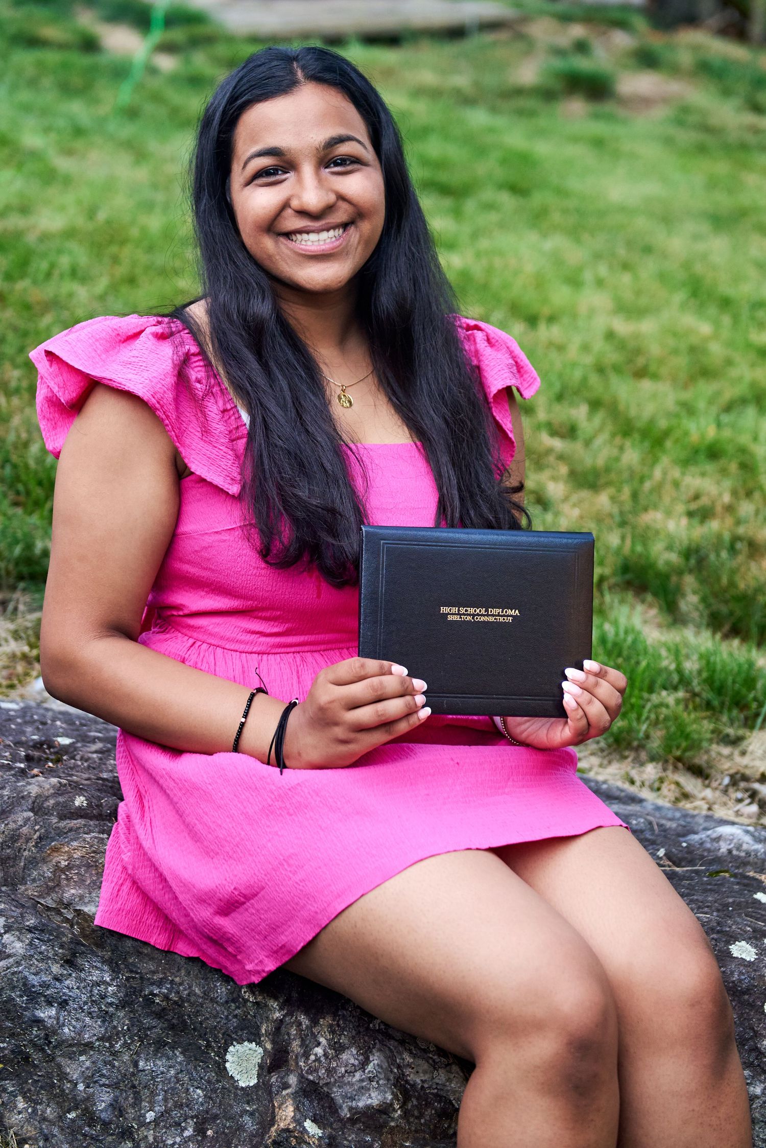 Person in bright pink top sitting outdoors holding a graduation diploma.