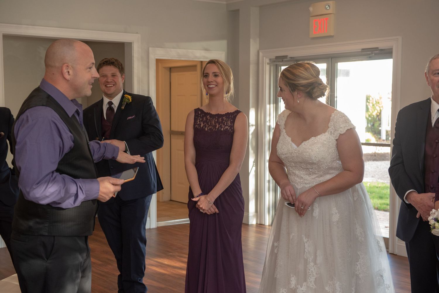 Formal wedding ceremony scene with attendees in purple and black attire in a sunlit room.