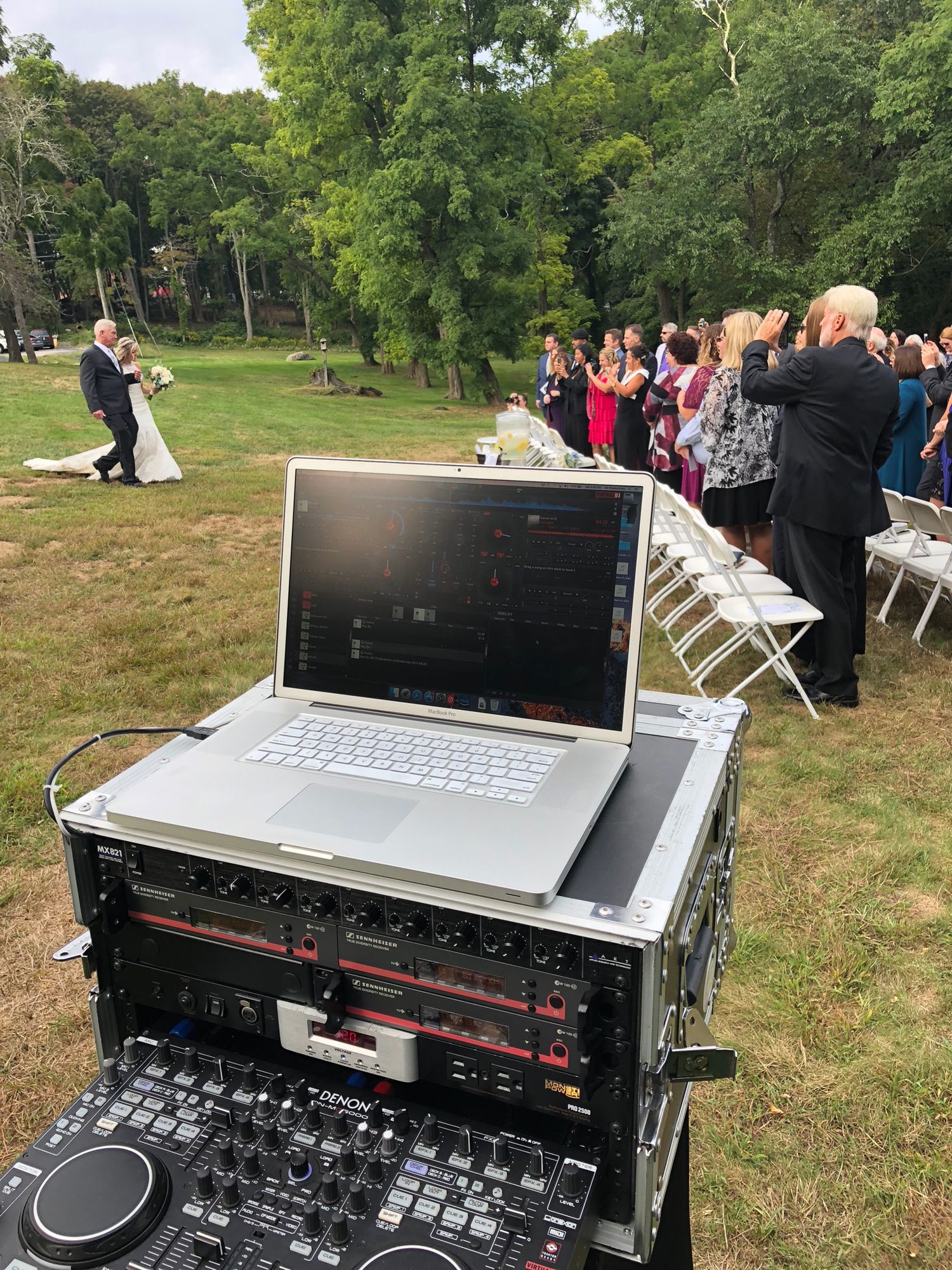 DJ equipment and laptop setup outdoors with wedding guests visible in the background.